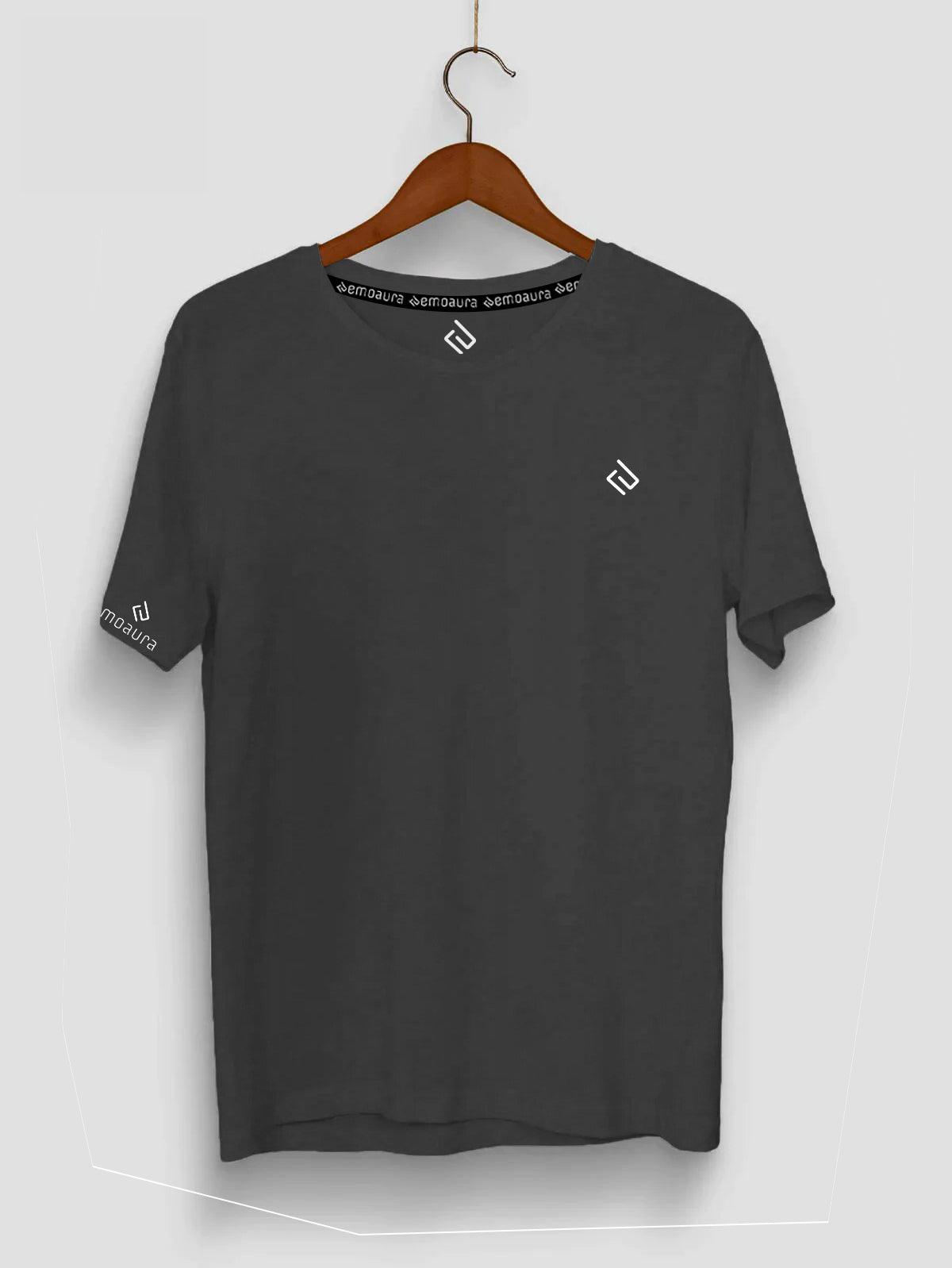 An "emoaura" Plain Classic Fit T-Shirt in dark grey blend cotton fabric, displayed on a wooden hanger. It features a small white geometric logo on the upper left chest, a matching logo on the left sleeve, and additional white text around the inside of the neckline. The background is plain white.